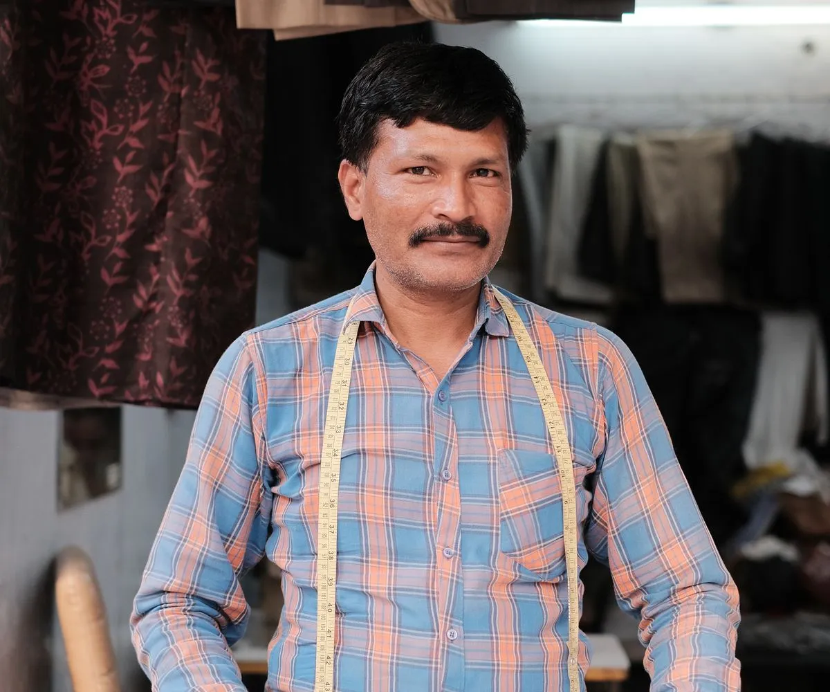 Tailor in his Lahore shop, tape measure around his neck, finished garments on the rail behind — silaaimarkaz